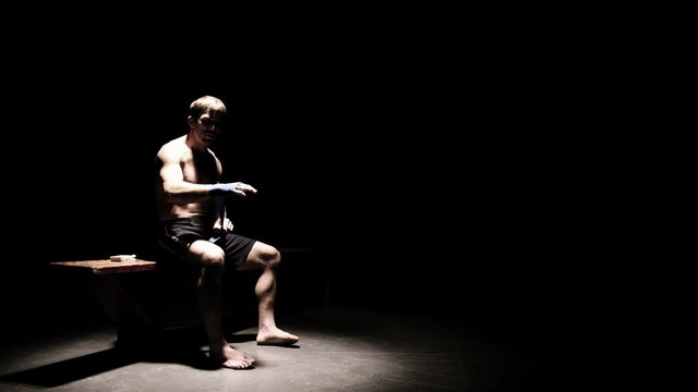 Athlete sitting on a bench wraps his hands. Wide shot.