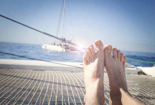 man lounging on a catamaran sailboat trampoline with his feet propped up and crossed. calm blue ocean and cloudless blue sky are in the background