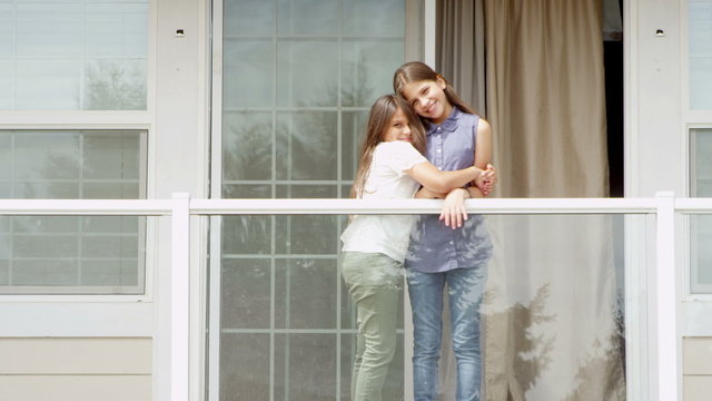 A young girl hugs her sister and they smile while standing on a balcony