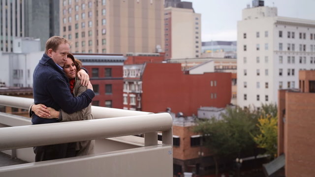 A couple embraces each other as they overlook the city from a tall building