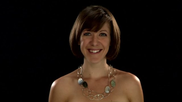 A woman with short hair wearing a statement necklace gestures the camera to come to her