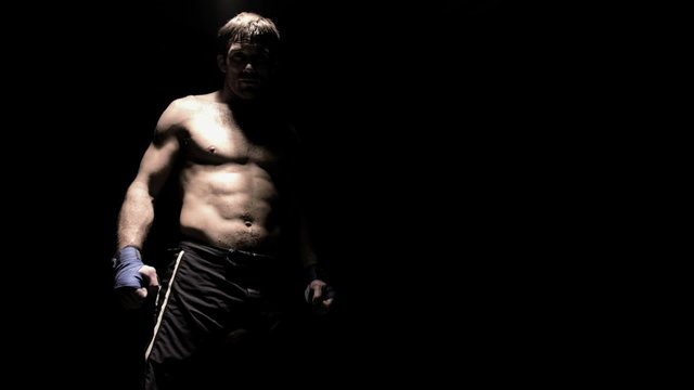 A fighter shadow boxes with the camera under a light in a dark room