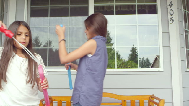 Two young girls stand on their porch and wave their bubble wands around