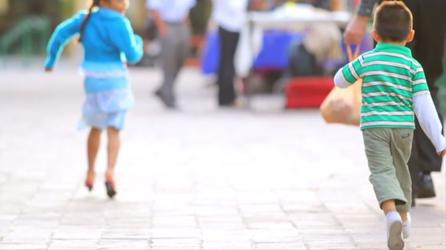 A little Latin girl and boy run down a street chasing each other