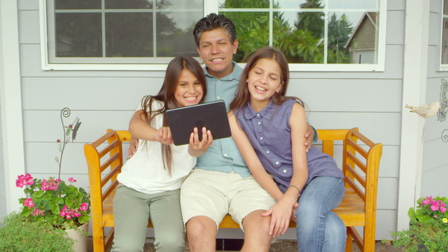 A father and his two daughters sit on a bench outside of their house and take pictures of themselves with a tablet