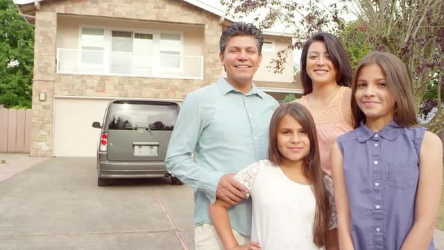 An hispanic family stand outside in their driveway looking and smiling at the camera