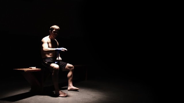 Athlete sitting on a bench wraps his hands. Wide shot.