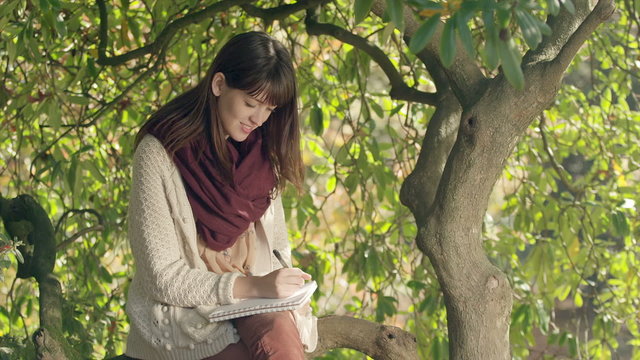 A young woman sits in a tree and writes in her notebook