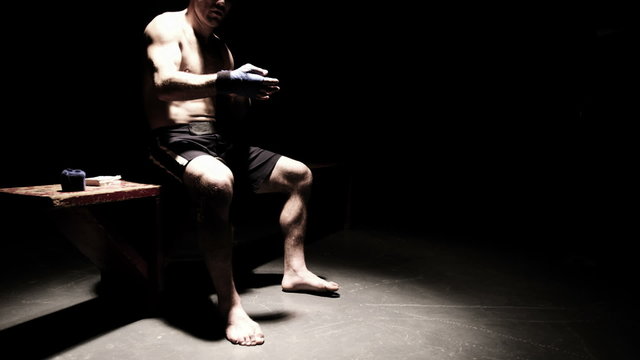 Mixed martial arts athlete sits on a bench and tapes his hands. Wide shot.