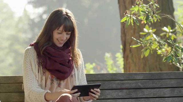 A girl sits on a bench in a park and takes pictures with her tablet
