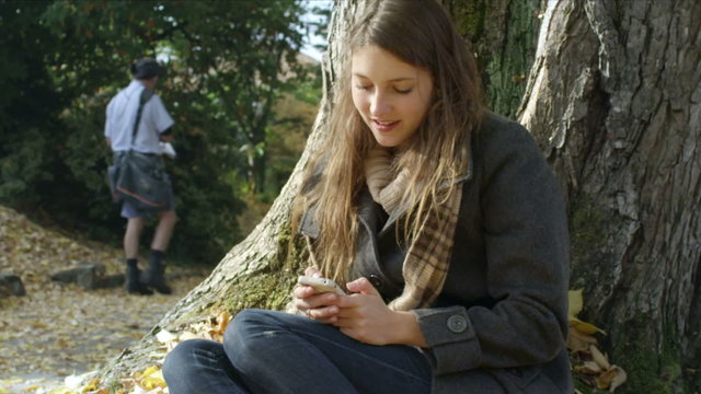 A young woman sits on a tree and texts on her phone while in the park
