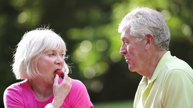 Wife feeds husband a strawberry during a picnic in a park. Medium shot.