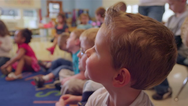 A group of young kids sit in on the floor and pay attention to their teacher