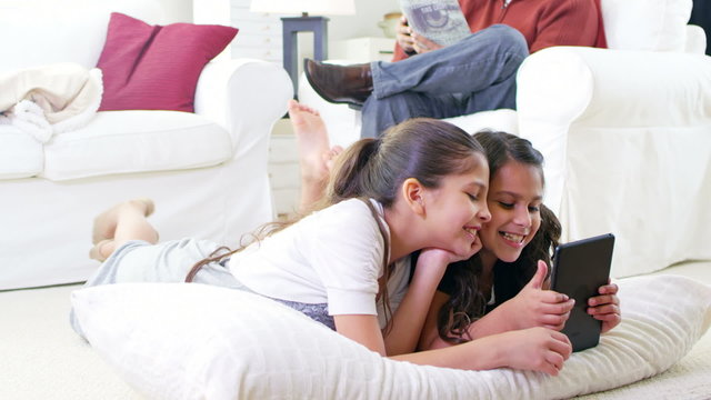 Two little girls lie on a pillow together and play with a tablet computer