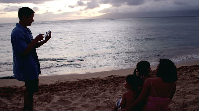 A man taking a picture of his family sitting in the sand at the beach at sunset