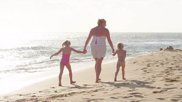 A mom and her two daughters hold hands and walk down the beach together