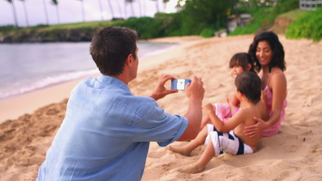 A man takes a picture of his family while they sit in the sand at the beach