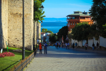 People walk along old street with the sea on background.