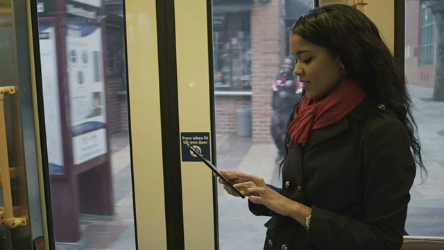 A girl stands up in the streetcar as it moves and she plays with her tablet