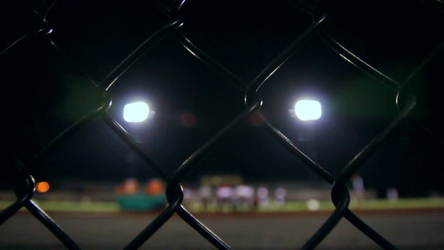 Dolly behind a fence viewing a high school football team as they warm up for a night time game.
