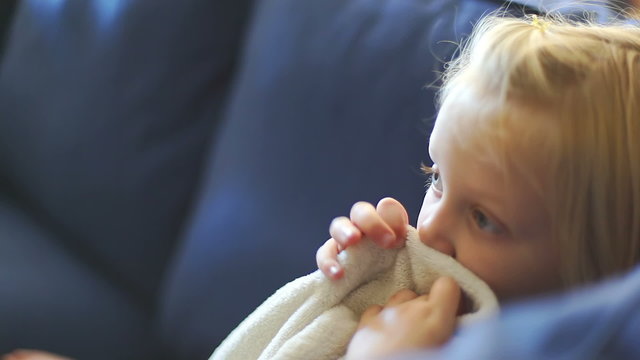 A young girl sits and holds her security blanket to her face and sucks her thumb