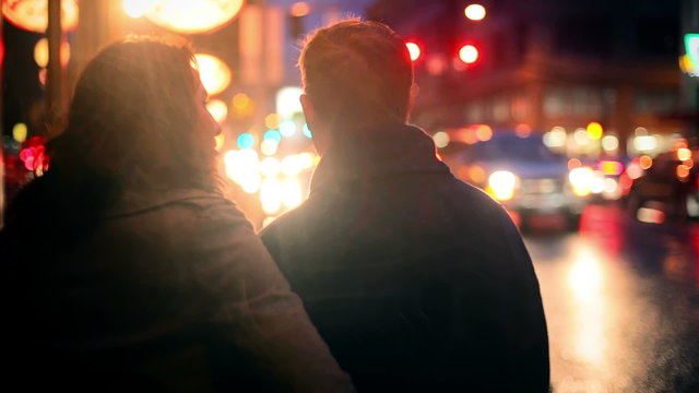 A Couple Walking Away From The Camera At Night, With Lots Of City Lights