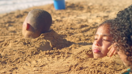 Young kids play at the beach and are buried up to their necks in sand