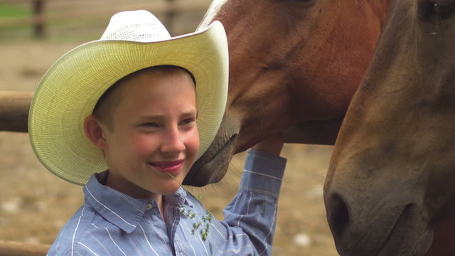 A boy in a cowboy hat stands in front of a horse with grass in his mouth