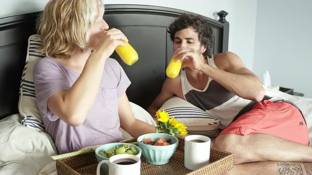 A man and woman lay in bed together drinking juice and kissing
