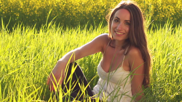 A cute young woman sits in the grass in an open field and looks at the camera