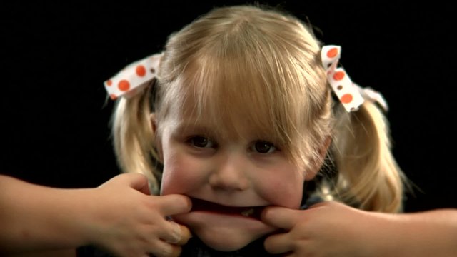 A  young blonde girl with pigtails pulls her mouth to make a funny face