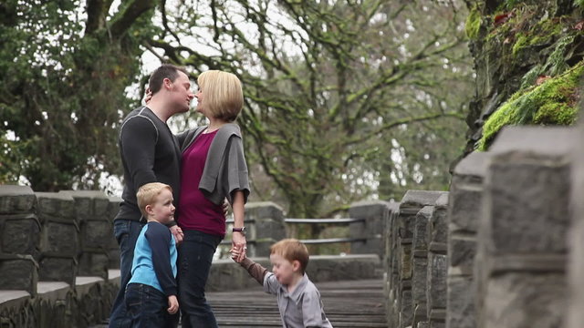 A mom and dad with their two kids walk down a set of stairs in a park and kiss