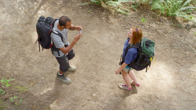 The camera looks down on a man who has stopped hiking the trail so he can take a picture of his girlfriend
