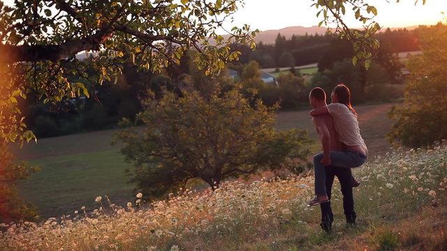 Boy gives girl piggyback ride in field during sunset. Wide shot.