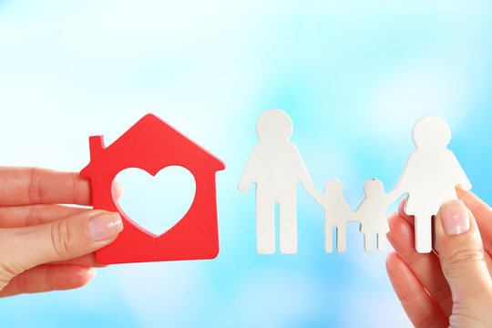Female hands holding model of house and paper family on bright background