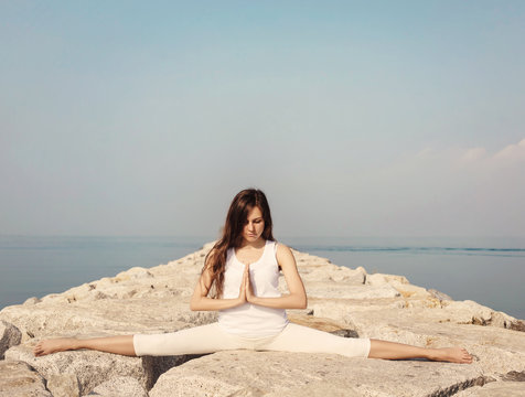 Beautiful young woman in white clothes practicing yoga on the beach on the rocks. Samakonasana pose