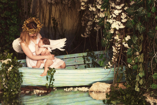 Young mother breastfeeding newborn baby dressed as angels with white wings in a boat on the river