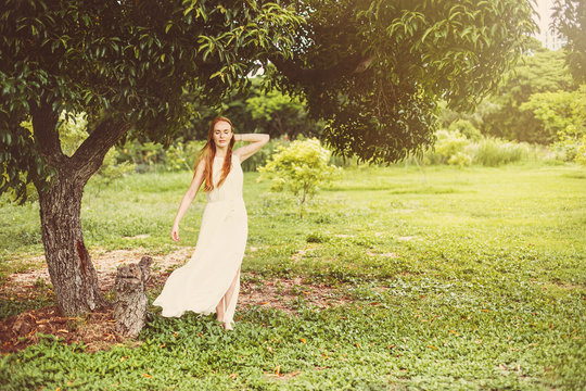 Beautiful young woman in long white dress with long red hair standing in wind under big tree on  green meadow