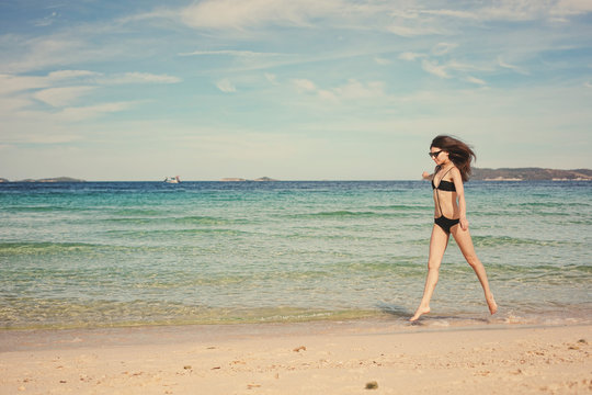 Beautiful happy young brunette woman in swimsuit running along the beach near blue sea