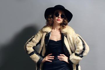 Portrait of beautiful model in fur coat, hat and sunglasses on gray background