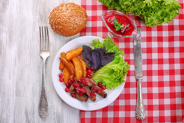 Beef with cranberry sauce, roasted potato slices, vegetables and bun on plate, on color wooden background