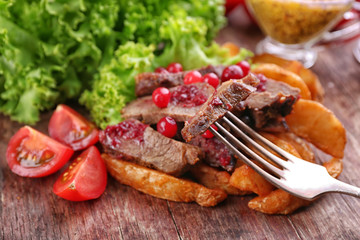 Beef with cranberry sauce, roasted potato slices and bun on cutting board, on wooden background