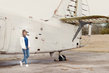 Beautiful girl in jeans and a white jacket standing near an old airplane on a cloudy day