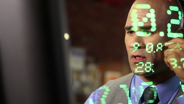 Anxious looking stock trader with ticker symbols projected across him.  Close up with dolly move past computer screen.  All ticker symbols are fictitious companies.