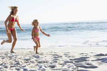 Mother And Daughter Running Along Beach Together Wearing Swimming Costume