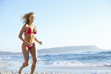 Obraz premium Young Woman Running Along Sandy Beach On Holiday Wearing Bikini