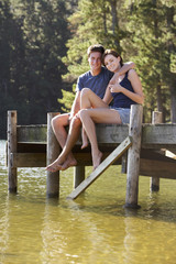 Young Romantic Couple Sitting On Wooden Jetty Looking Out Over Lake
