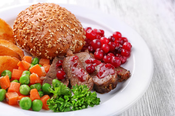 Beef with cranberry sauce, roasted potato slices, vegetables and bun on plate, on color wooden background