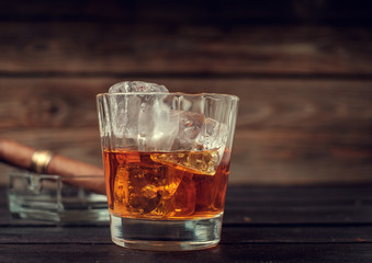 Glass of whiskey with ice and cigar on a wooden background.