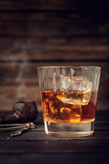 Glass of whiskey with ice and pipe on a wooden background.